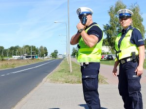 Policjant przykładając urządzenie do oka sprawdza prędkość. Po jego lewej stronie stoi policjantka.