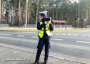 Na zdjęciu policjantka w maseczce dokonuje pomiaru prędkości pojazdu przy użyciu ręcznego miernika prędkości.