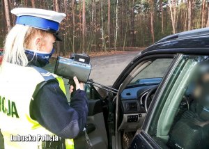 Na zdjęciu policjantka w maseczce pokazuje zatrzymanemu do kontroli drogowej mężczyźnie ręczny miernik prędkości, na którym widnieje przekroczona dopuszczalna prędkość.