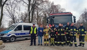 Policjanci i strażacy na tle policyjnego radiowozu.