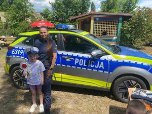 Policjantka z dziewczynką, która trzyma tarczę do zatrzymywania pojazdów, na tle radiowozu.